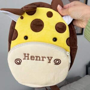 May include: A yellow and brown plush giraffe backpack with a zipped pocket and adjustable straps. The backpack has a white embroidered patch with the name "Henry" on it.
