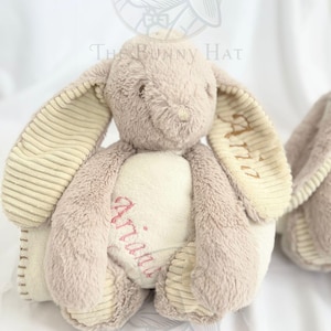 May include: A plush bunny rabbit in soft beige and cream colors. The bunny has long, floppy ears with corduroy-like texture on the inside. The name "Ariana" is embroidered in pink on the bunny's chest. The Bunny Hat logo is visible in the background.