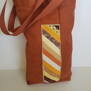 Peut inclure: Un sac fourre-tout en toile marron avec un motif patchwork sur le devant. Le patchwork est composé de tissus de différentes couleurs, dont le jaune, l'orange, le marron et le blanc. Le sac a deux longues anses.