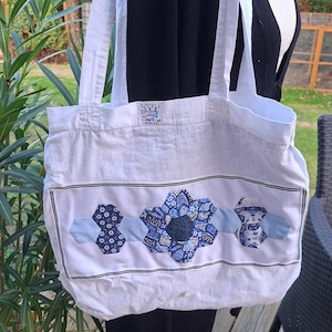 Op de afbeelding: Een witte tote bag met twee lange schouderbanden en een centraal horizontaal paneel versierd met blauwe en witte stoffen zeshoeken in bloemen- en geometrische patronen. Een klein rechthoekig label bevat de tekst Les points mêlés.