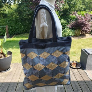 Peut inclure: Un grand sac fourre-tout avec un motif géométrique. Le sac présente un design patchwork avec du tissu bleu, beige et à motifs. Il a un dessus zippé et des poignées robustes. Le sac est présenté à l'extérieur.