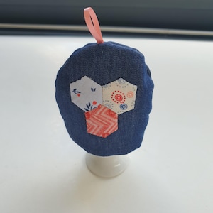 Peut inclure: Un protège-œuf bleu denim avec trois patchs hexagonaux décoratifs à motifs floraux et géométriques. Une boucle de ruban rose est attachée en haut. Le protège-œuf est présenté sur un coquetier en céramique blanche.