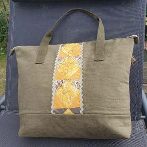 Peut inclure: Un grand sac fourre-tout en toile vert olive avec une fermeture éclair. Le sac a un design patchwork sur le devant, avec une variété de carrés de tissu jaune, orange et marron.