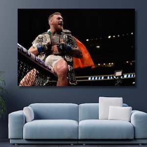 May include: A man in a black and green robe with an orange sash is celebrating in a boxing ring. He is wearing white shorts and blue gloves. The ring is surrounded by a black fence and there are lights in the background.