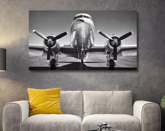 May include: A black and white wall art print featuring a front-facing view of a vintage twin-engine propeller airplane on a runway. The aircraft displays a metallic finish, two large propellers, and landing gear, captured in a high-contrast monochrome.