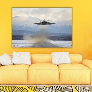 Puede incluir: Un cuadro rectangular que muestra un avión de combate F-22 Raptor gris despegando de una pista cubierta de nieve. La aeronave está centrada en el aire con una estela de humo debajo, sobre un fondo de montañas lejanas y nubladas.