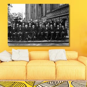 May include: A black and white rectangular wall art print featuring a large group of men in formal suits posing for a photograph in front of a stone building. The print is displayed above a bright yellow sofa with white cushions and a yellow rug.