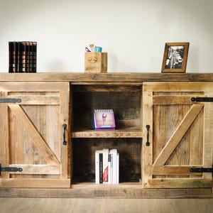 Könnte beinhalten: Ein rustikaler Holzschrank mit Schiebetüren und einem offenen Mittelfach. Der Schrank hat eine natürliche Holzoberfläche, schwarze Beschläge und ein gerahmtes Foto. Ein Kalender, Bücher und Bürobedarf sind sichtbar.