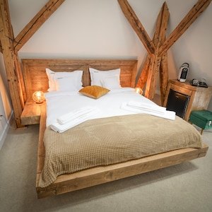 Peut inclure: Un lit avec un cadre en bois, une couverture beige et des oreillers blancs. Le lit est dans une pièce avec des poutres en bois apparentes et une petite table de chevet en bois avec une lampe.