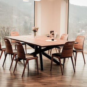 Könnte beinhalten: Ein Esstisch aus Holz mit einem schwarzen Metallgestell und sechs Stühlen. Der Tisch ist mit einem Glas Wein und einer Flasche Wein gedeckt. Der Tisch steht vor einem großen Fenster mit Blick auf einen Wald.