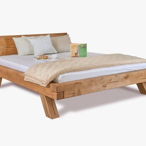 Könnte beinhalten: Ein rustikaler Holzbettenrahmen mit Kopfteil, mit einer hellen Matratze, Kissen und einer beige gesteppten Decke. Ein Buch und ein Croissant liegen auf dem Bett. Der Bettrahmen besteht aus dickem, unbehandeltem Holz.