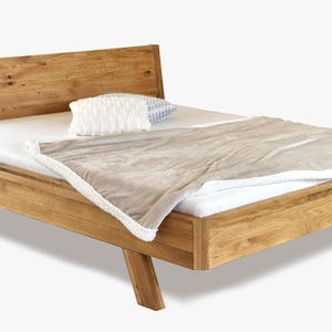 Pode incluir: Cama de madeira com cabeceira, colchão branco, cobertor bege com acabamento branco e uma almofada decorativa. A estrutura da cama é feita de madeira clara. A cama tem cerca de 200 cm de comprimento.