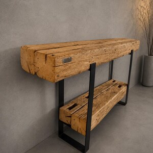 Peut inclure: Une console composée de deux poutres en bois clair patiné, empilées horizontalement pour former le plateau et l'étagère inférieure, soutenues par une structure en métal noir. Une petite plaque métallique est fixée sur la poutre supérieure.
