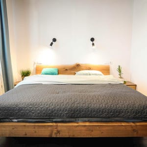 Peut inclure: Une couette matelassée grise sur un cadre de lit en bois avec une tête de lit en bois massif. Deux appliques murales avec des luminaires noirs sont fixées au mur derrière le lit.