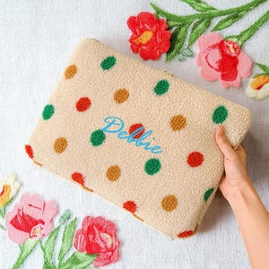 Puede incluir: Una funda beige para portátil con un estampado de lunares rojos, verdes y marrones. El nombre "Debbie" está bordado en azul. La funda es rectangular y tiene un aspecto suave y texturizado.
