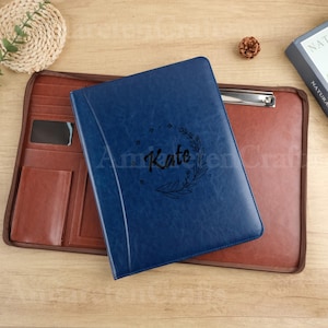 May include: A blue leather portfolio with the name "Kate" and a floral design. The portfolio is on top of a brown leather document holder with pockets and a clipboard. The background is a wooden surface.