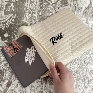 Puede incluir: Funda acolchada para tableta de color crema con cierre de cremallera. La palabra "Rose" está escrita en escritura negra en la parte delantera. Una tableta con pegatinas es parcialmente visible dentro.