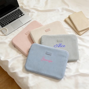 May include: Four pastel-colored laptop sleeves, including pink, cream, and light blue, are arranged on a white satin surface. Each sleeve has a rectangular label and a zipper closure. Two sleeves are personalized with names in cursive.