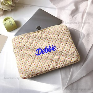 May include: A quilted laptop sleeve with a cream background and a pattern of small red hearts and yellow flowers. The name "Debbie" is written in blue cursive on the front. A silver laptop is partially visible inside the sleeve.