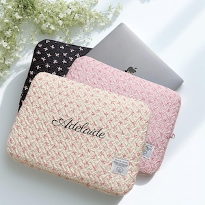 Puede incluir: Tres fundas para portátiles en negro, rosa y crema, con un patrón texturizado. La funda crema tiene el nombre "Adelaide" bordado. Un portátil plateado es parcialmente visible.