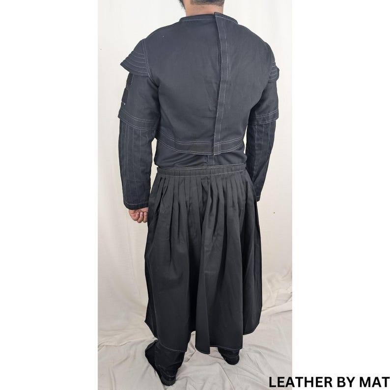 K&ouml;nnte beinhalten: Ein schwarzes Langarmhemd mit einem schwarzen Rock. Das Hemd hat ein wei&szlig;es N&auml;hdetail am Kragen und an den Schultern. Der Rock hat ein Faltendesign und wei&szlig;e N&auml;hte am Saum.