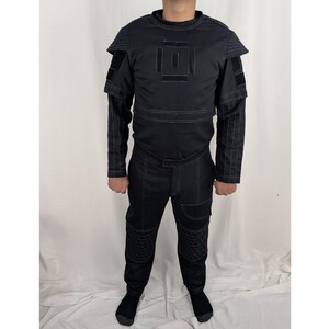 K&ouml;nnte beinhalten: Ein schwarzer Overall mit geometrischem Muster auf der Brust und Schulterpolstern. Der Overall hat einen Rei&szlig;verschluss und einen G&uuml;rtel.