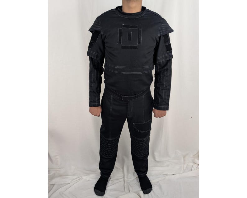 K&ouml;nnte beinhalten: Ein schwarzer Overall mit geometrischem Muster auf der Brust und Schulterpolstern. Der Overall hat einen Rei&szlig;verschluss und einen G&uuml;rtel.