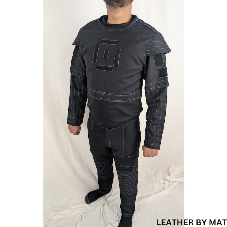 K&ouml;nnte beinhalten: Ein schwarzer, lang&auml;rmliger Overall mit geometrischem Design auf der Brust und gesteppten Details an den Beinen. Der Overall hat einen hohen Kragen und einen Rei&szlig;verschluss. "LEATHER BY MAT" ist in der unteren rechten Ecke des Bildes gedruckt.