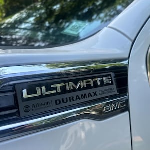 May include: Close-up of a white GMC truck's front end, showcasing a black and silver "ULTIMATE DURAMAX" emblem. The emblem includes "Allison Transmission" and "Turbo Diesel" text. The vehicle's chrome trim and a portion of the windshield are visible.