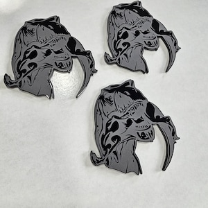 May include: Three gray and black enamel pins featuring a stylized monster head with large claws. The pins are arranged on a white surface. The design has intricate details and a dark, ominous aesthetic.