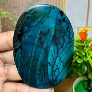 May include: Oval-shaped gemstone with a striking pattern of teal and dark blue veins. The polished surface has a glossy finish, with a dark, almost black, border. The stone's design resembles a cracked or fractured texture, creating a unique visual effect.