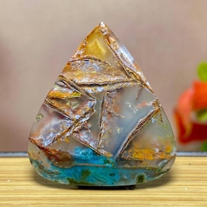 Peut inclure: Une pierre précieuse en forme de goutte d'eau avec une mosaïque de couleurs. Le sommet est d'un jaune-brun chaud, passant à un mélange de blanc, d'orange et de brun. La base présente une teinte turquoise vibrante, créant un contraste saisissant. La surface de la pierre est polie et brillante.