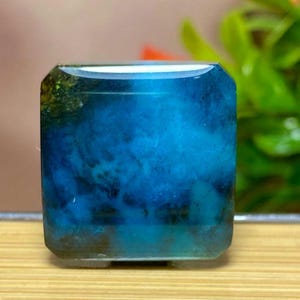 May include: A square, polished gemstone with a captivating blue and teal color palette. The stone's interior features a cloudy, marbled effect, with darker shades of blue and hints of green. The gemstone is set on a wooden surface.