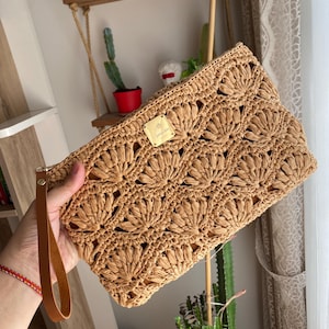 Könnte beinhalten: Eine hellbraune gewebte Clutch mit einem Muschelmuster. Die Clutch hat eine braune Leder-Handschlaufe und ein kleines goldfarbenes Metallschild. Die Tasche wird von einer Person gehalten.