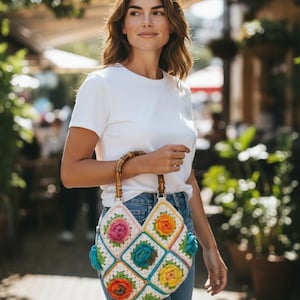 May include: A white crochet bag with a bamboo handle, featuring colorful floral designs. The bag has a patchwork pattern with squares of pink, orange, yellow, and blue flowers. The bag is held by a person wearing a white t-shirt and blue jeans.