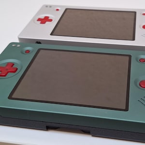 Può includere: Due console di gioco portatili con schermo rettangolare, un pad direzionale rosso e pulsanti azione rossi. Una console ha una finitura argento opaco, mentre l'altra è verde bosco con una base grigio scuro e un jack per cuffie laterale.