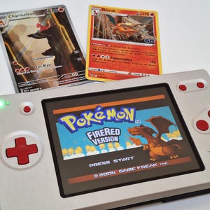 Peut inclure: Une console de jeu portable argentée avec des boutons et une croix directionnelle rouges, affichant l'écran titre de Pokémon FireRed Version. Deux cartes à collectionner Pokémon, Charmeleon et Glurak, sont placées derrière la console.