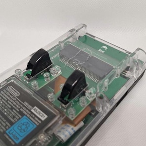 Könnte beinhalten: Ein transparentes Kunststoffgehäuse für elektronische Geräte mit sichtbaren internen Komponenten. Das Gerät enthält eine grüne Leiterplatte, einen Nintendo-Akku und schwarze Komponenten. Das Gehäuse hat eine rechteckige Form und ist teilweise transparent.