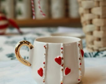 May include: A white ceramic mug with a gold handle and red heart accents. The mug has vertical beaded lines with red and gold details. The mug is on a white and blue patterned surface.