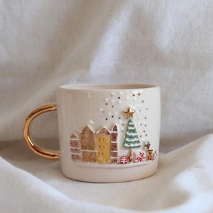 May include: A cream ceramic mug with a gold handle. The mug has an embossed festive design with buildings, a Christmas tree, and gifts. Gold star and dot accents add sparkle to the holiday-themed mug. The mug is approximately 9 cm tall.