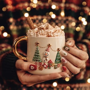 May include: A cream-colored ceramic mug with a gold handle, featuring a raised relief design of two green Christmas trees, a snowman, and colorful gift boxes. The mug is filled with whipped cream and a cinnamon stick.