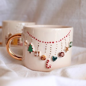 Puede incluir: Taza de cerámica color crema con asa dorada y adornos festivos. La taza presenta una guirnalda de puntos rojos y adornos colgantes, incluyendo un árbol de Navidad, regalos y bastones de caramelo. Una segunda taza es visible en el fondo.