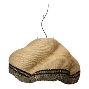 K&ouml;nnte beinhalten: Gewebte Pendelleuchte in Naturbeige mit schwarzem Band am unteren Rand. Der Schirm hat eine unregelm&auml;&szlig;ige Form und wird von einem schwarzen Kabel gehalten.