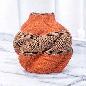 May include: A handwoven orange and brown basket with a unique, textured design. The basket features a cylindrical neck and a bulbous body with a woven pattern of diamonds and stripes. The basket is made of natural fibers.