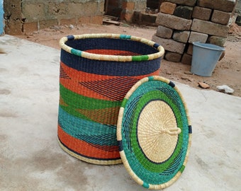 May include: A woven storage basket with a lid. The basket is made from natural materials and has a colourful striped pattern in shades of orange, green, blue, and black. The lid is also woven and has a similar pattern.