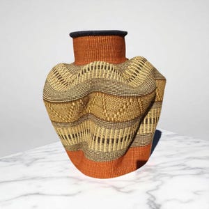 May include: Woven basket vase with a unique, textured design. The vase features a combination of orange, tan, and black woven patterns. The top rim is dark blue, and the base is orange. The vase is sitting on a white marble surface.