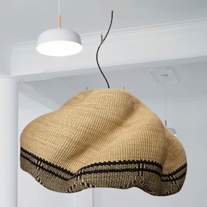 K&ouml;nnte beinhalten: Eine gro&szlig;e, gewebte Pendelleuchte in Naturbeige mit einem schwarzen geometrischen Muster am unteren Rand. Die Leuchte hat eine einzigartige, organische Form und h&auml;ngt an einem schwarzen Kabel. Im Hintergrund ist eine wei&szlig;e, runde Deckenleuchte zu sehen.