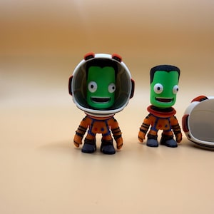May include: Two green astronaut figurines in orange and blue spacesuits and black boots. One figure wears a helmet, the other does not. The helmets are white with red detailing. The background is a plain light orange.