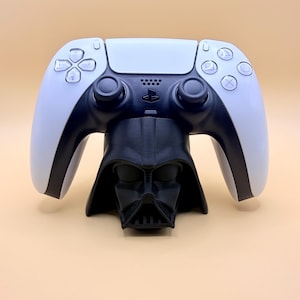 May include: A white and black PlayStation 5 controller sits on a black Darth Vader-themed stand. The controller has a central black section with white side panels and button details. The stand is a detailed bust of Darth Vader.