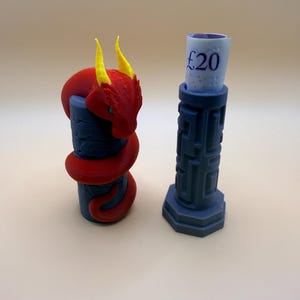 May include: Two 3D-printed money holders. One is a blue cylinder with a red dragon wrapped around it, featuring yellow horns. The other is a blue, geometric pillar holding a rolled-up £20 note. The background is a solid, light beige.